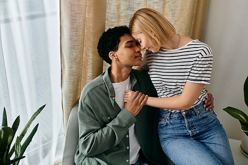 A young multicultural couple sits together, deeply connected, in a modern apartment with large windows and soft lighting.