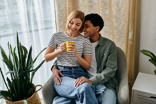 A young, interracial couple shares a warm drink while relaxing in a comfortable, modern apartment.