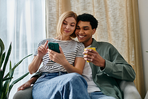 A young multicultural couple enjoys a relaxing afternoon in their modern apartment, looking at a phone and sharing a cup of tea.