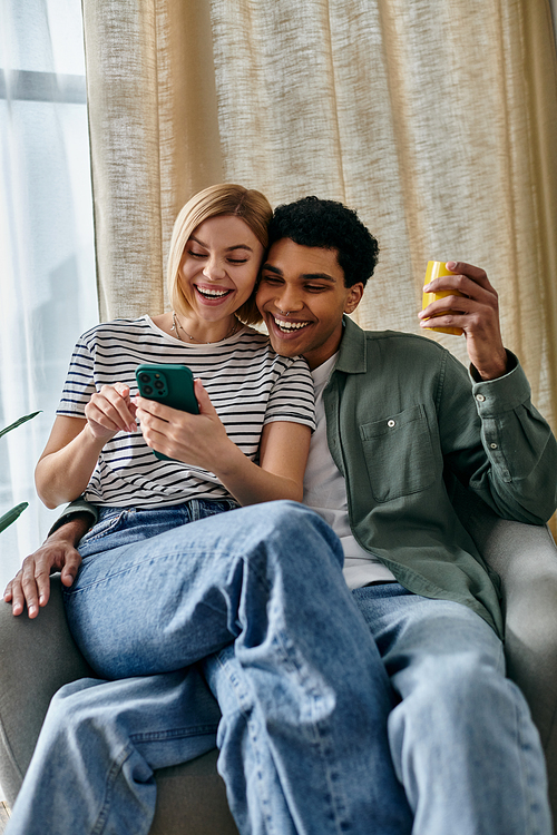 A young interracial couple share a laugh while looking at their phone in their modern apartment.