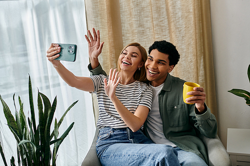 A young, multicultural couple smiles for a selfie in their modern apartment, showcasing a relaxed and intimate moment.