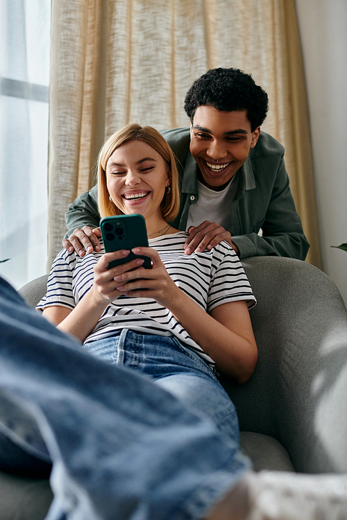 A young multicultural couple relaxes on a modern couch in their apartment, laughing together while looking at a smartphone.