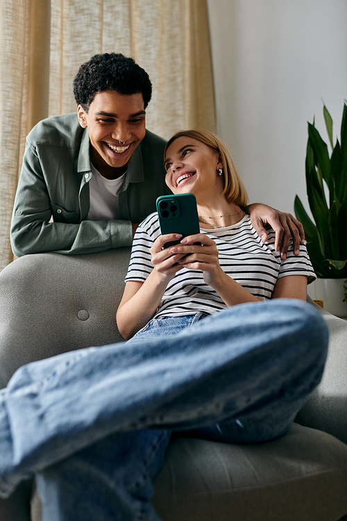 A young multicultural couple relaxes on a couch in a modern apartment, laughing and looking at a smartphone.