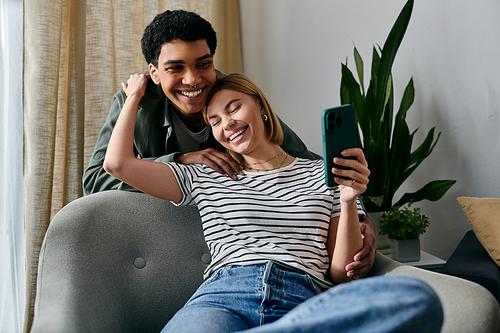 A young interracial couple laughs together in a modern apartment while holding a smartphone.