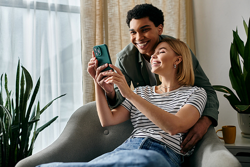 A young interracial couple happily looks at a smartphone together while relaxing on a couch in their modern apartment.