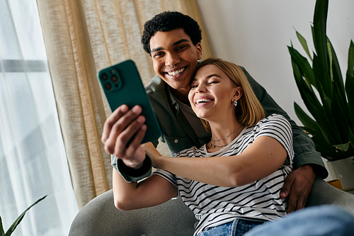 A young multiracial couple happily takes a selfie in a modern apartment, smiling and enjoying each others company.