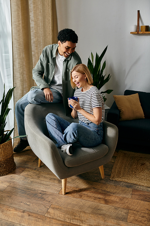 Young couple enjoying each others company in a modern apartment. Woman sits in gray armchair as man leans against back of it