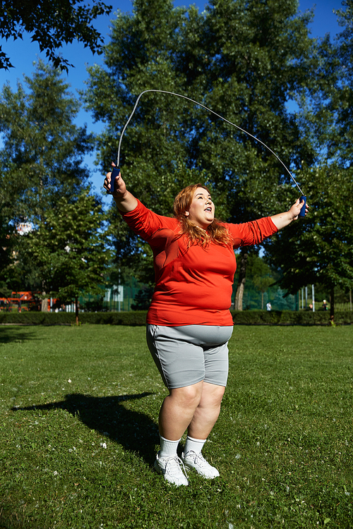 A plus size woman joyfully skips rope in a vibrant park.