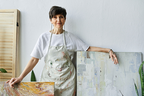 A woman in an apron stands proudly by her canvases in a sunlit art studio.