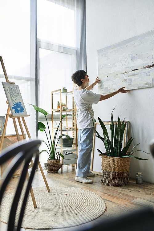 A mature woman curates her art in a sunlit studio filled with plants.