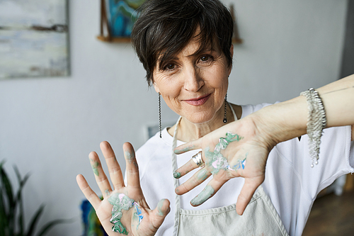 A mature woman joyfully showcases her paint splattered hands while working.