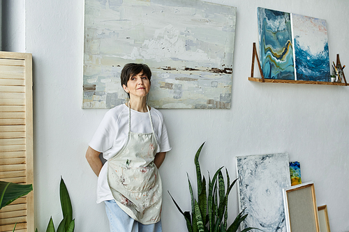 A mature woman stands in her art studio, contemplating her colorful paintings.