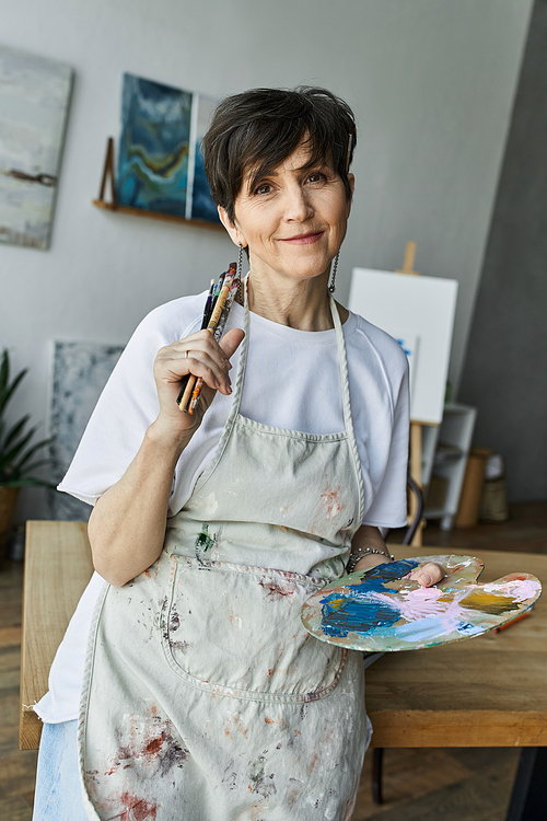 A woman in an art studio smiles while holding brushes and a colorful palette.