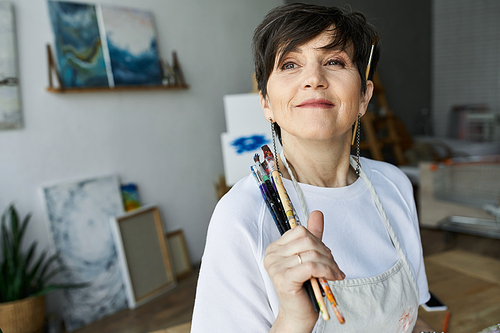 A painter smiles, holding brushes in her vibrant art studio.