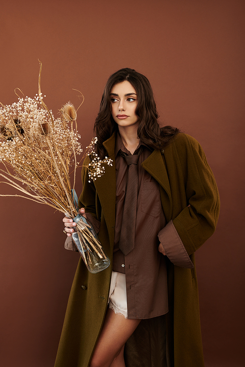 A young woman poses elegantly in a stylish autumn look, holding dried flowers against a warm backdrop.