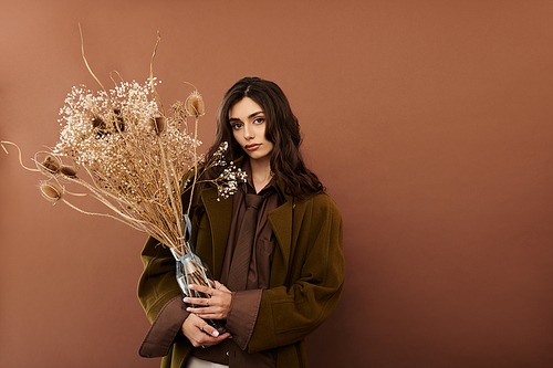 A stylish young woman elegantly poses in autumn attire, holding a bouquet of dried flowers against a warm backdrop.