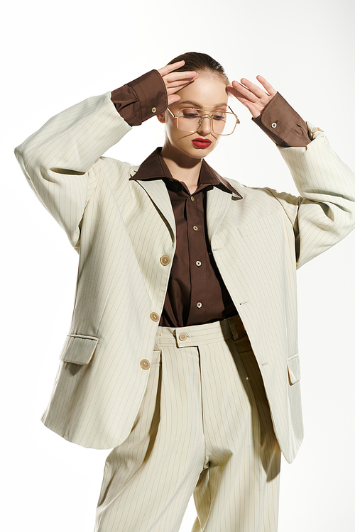 A woman poses wearing a beige suit, brown shirt, and gold-rimmed glasses.