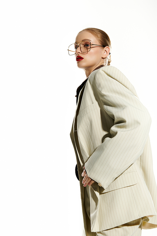 A woman wearing a white blazer and large glasses poses against a white backdrop.