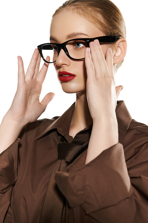 Woman adjusts stylish glasses, red lips adding color to elegant brown attire.