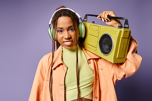 happy african american woman in wireless headphones standing with retro boombox on purple backdrop