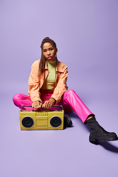 stylish african american woman with dreadlocks sitting next to retro boombox on purple backdrop