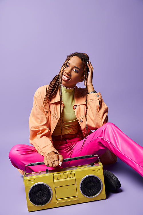 happy african american woman with dreadlocks sitting next to retro boombox on purple backdrop