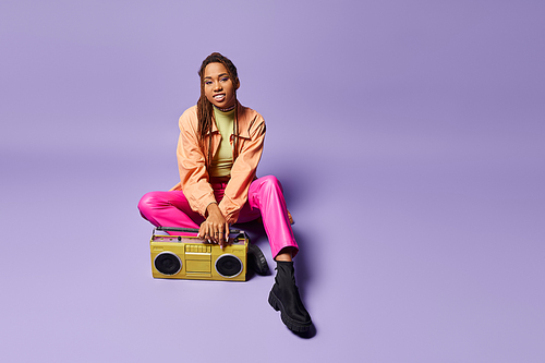 cheerful african american woman with dreadlocks sitting next to retro boombox on purple backdrop