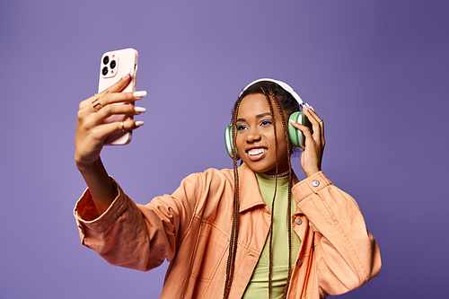 Cheerful african american woman in 20s taking selfie with her headphones on lilac backdrop