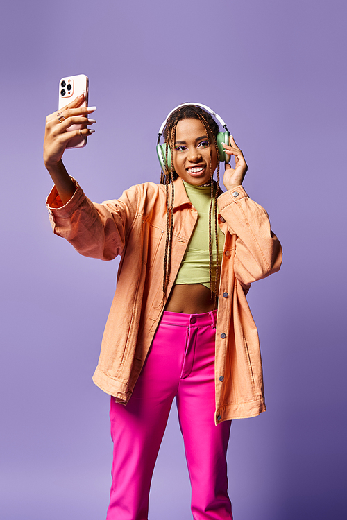 happy african american woman in 20s taking selfie in her wireless headphones on lilac backdrop