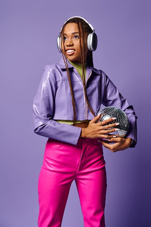 smiling young african american woman in stylish attire and headphones holding disco ball on purple