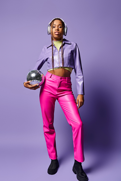 young african american woman in stylish attire and headphones holding disco ball on purple backdrop