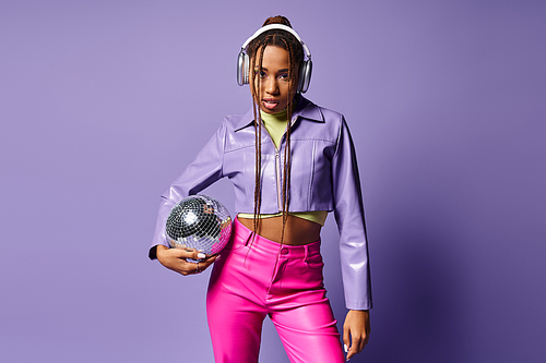 young african american woman in trendy attire and headphones holding disco ball on purple backdrop