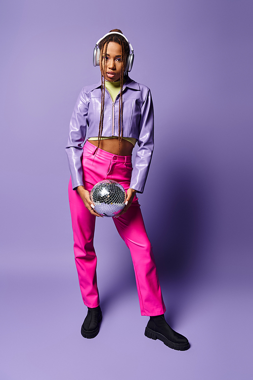 young african american woman in stylish wireless headphones holding disco ball on purple backdrop