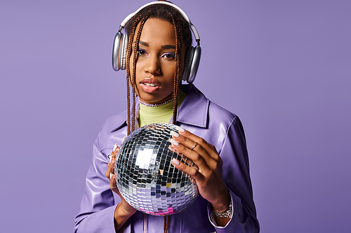 young african american girl in stylish wireless headphones holding disco ball on purple backdrop