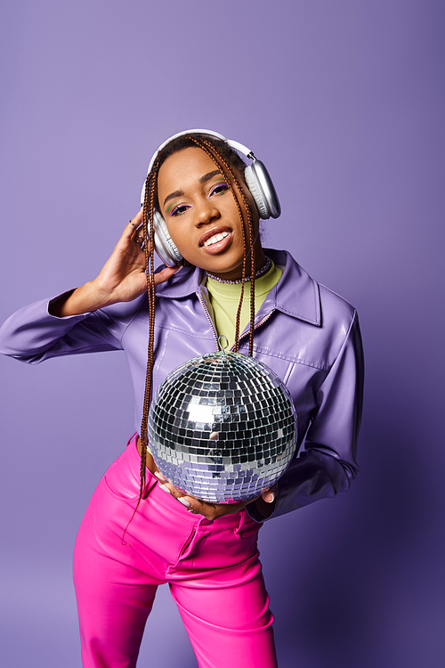 happy young african american woman in stylish attire and wireless headphones holding disco ball