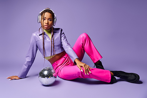 stylish african american girl in wireless headphones sitting near disco ball on purple backdrop