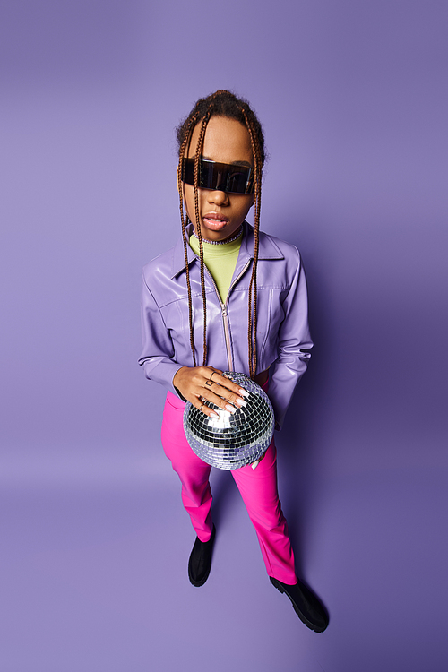 young african american woman in trendy sunglasses holding disco ball on chain and standing on purple