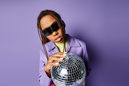 portrait of african american woman in trendy sunglasses holding disco ball on on purple background