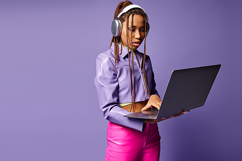 Focused african american woman with headphones working on laptop remotely on purple background