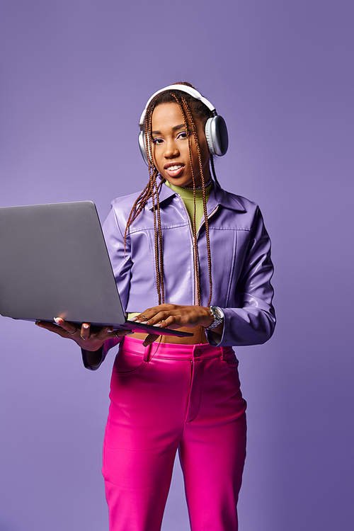 young african american woman with headphones working on laptop remotely on purple background