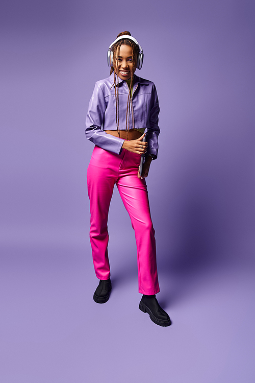 happy african american woman with headphones holding laptop and standing on purple background