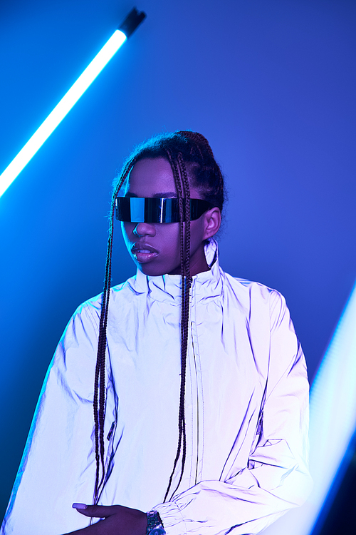 african american girl with dreadlocks posing in trendy jacket and sunglasses on blue with neon light