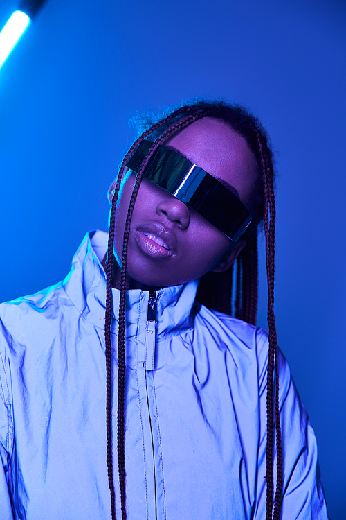 african american girl with dreadlocks posing in jacket and trendy sunglasses on blue with neon light