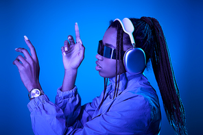 dreamy african american woman posing in trendy sunglasses with headphones on blue with neon light