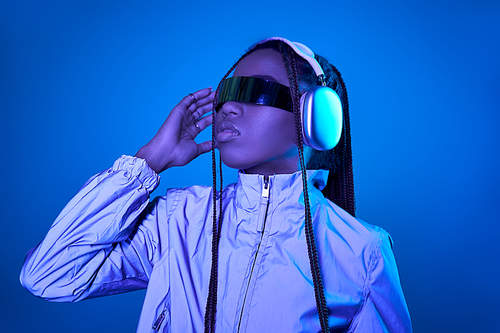 pretty dark skinned woman posing in trendy sunglasses with headphones on blue with neon light