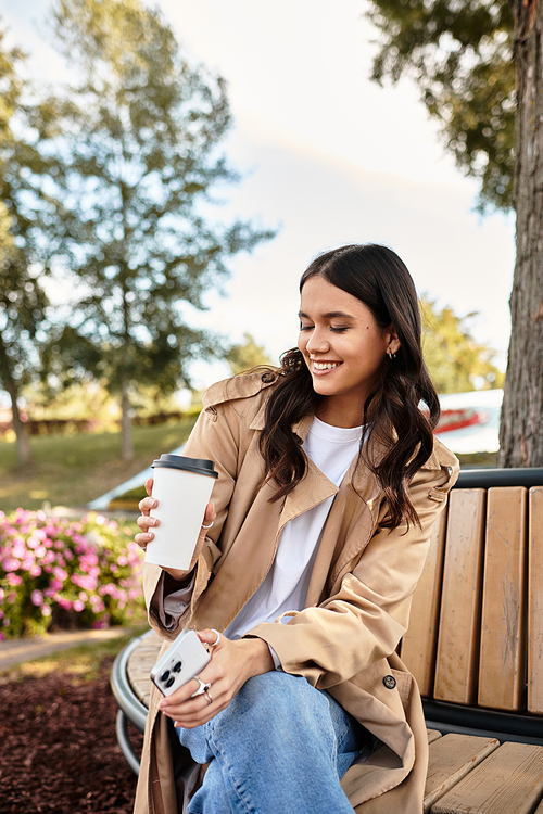 Smiling young woman dressed for autumn sits on a park bench, savoring her warm beverage.