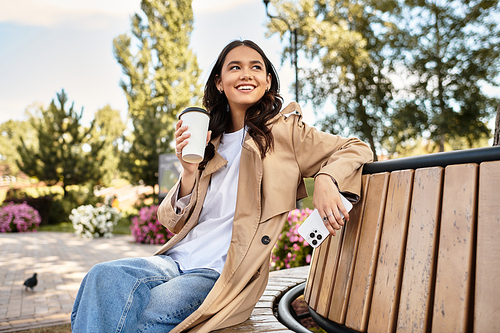 In a colorful park, a young woman dressed for autumn leisurely sips coffee while smiling.