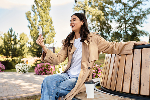 A cheerful young woman dressed warmly sits on a bench, capturing the beauty of fall with her phone.