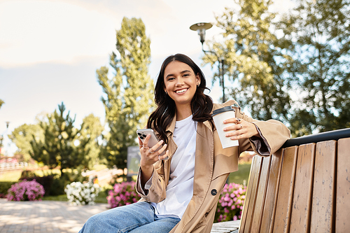 A cheerful woman enjoys coffee and texts on a park bench during autumn.