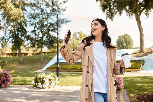 A joyful woman dressed for autumn strolls through a vibrant park, sipping coffee and smiling.